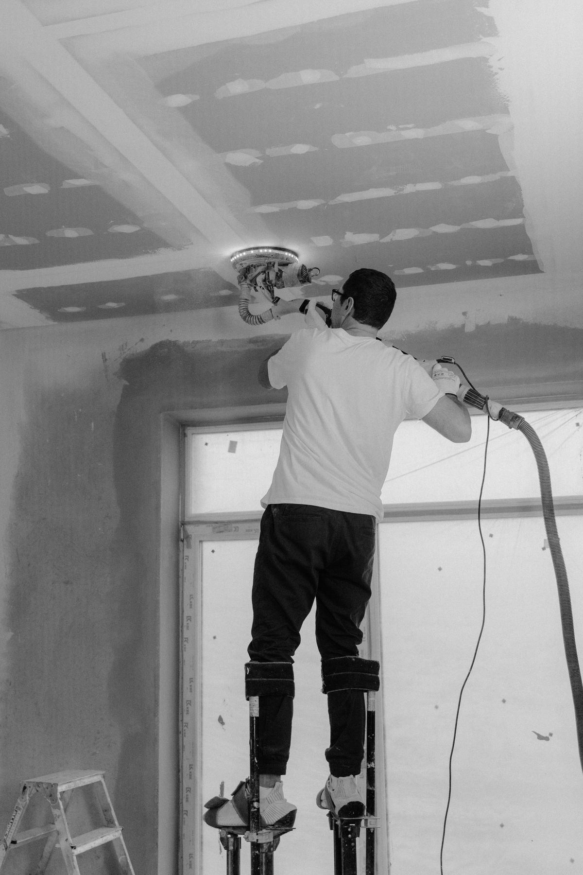 Worker on drywall stilts running a sander with dust collection across a taped ceiling