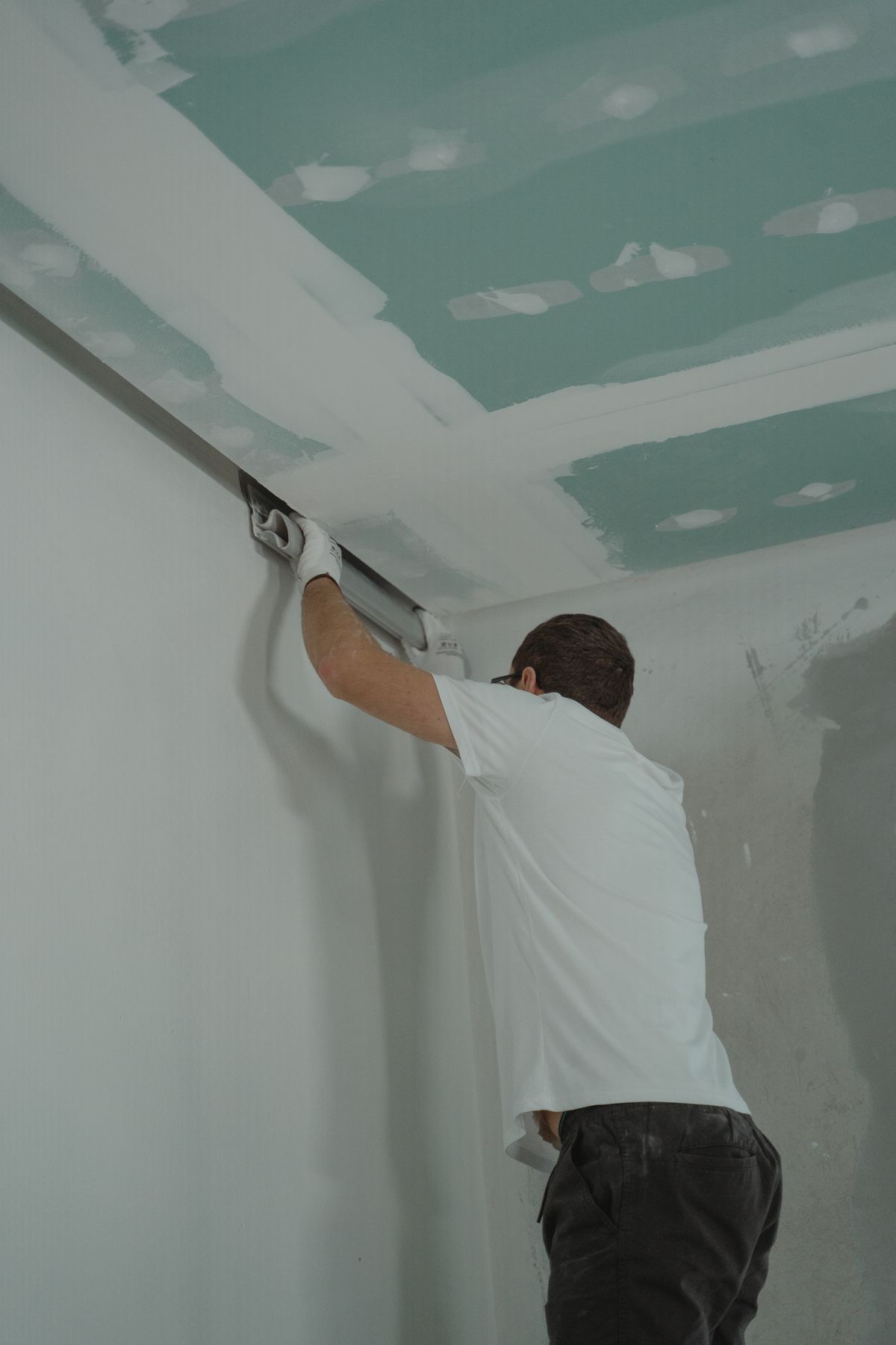 Tradesman finishing the ceiling-to-wall corner of a drywalled room with a wide knife